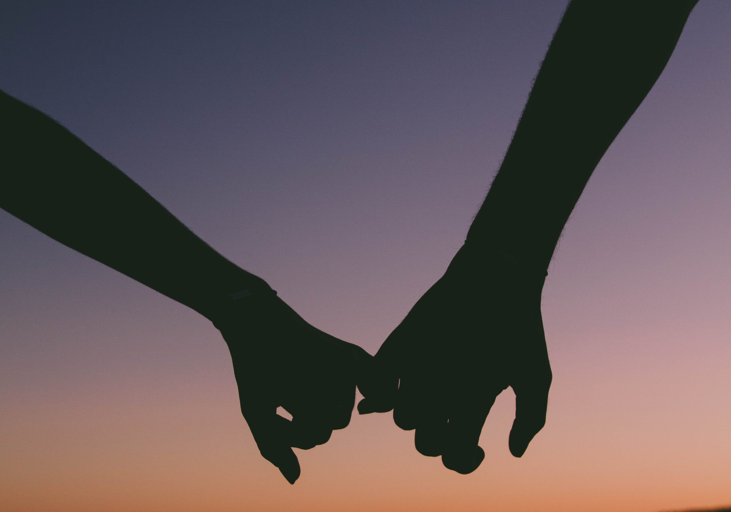 A silhouette of a man and woman intertwining pinkies against a beautiful sunset.